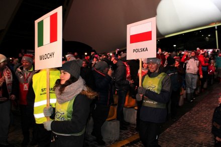 Wolontariusze z tablicami prezentującymi kraje uczestniczące w Pucharze Świata w Wiśle idą przez widownię amfiteatru